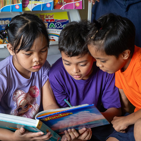 three children wearing purple and orange shirts reading a book together