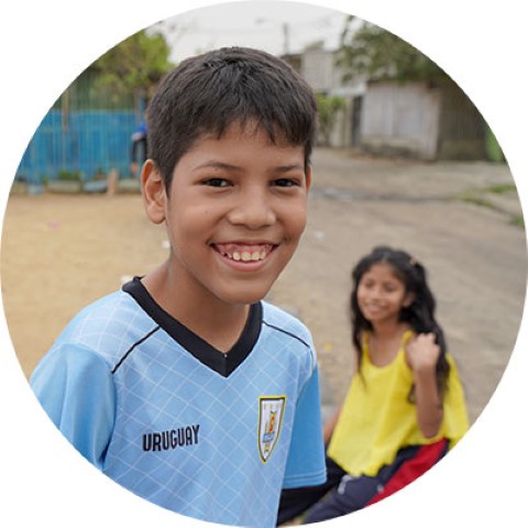 A boy smiles for the camera, a girl sits in the background